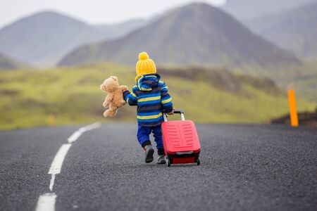 Cute toddler boy with teddy bearand suitcase in hand, running on a road in Iceland on a rainy dayの写真素材