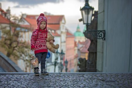 Beautiful toddler child with lantern and teddy bear, casually dressed, looking at night view of Prague city, wintertimeの写真素材