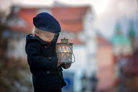Beautiful toddler child with lantern and teddy bear, casually dressed, looking at night view of Prague city, wintertimeの写真素材