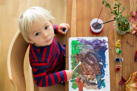 Sweet toddler boy with red apron, painting at home with water colors