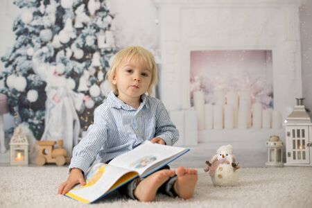 Blond toddler child, boy, reading a book at home, lying on the floor with a toyの写真素材