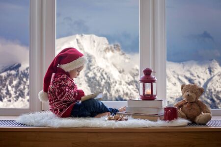 Sweet blonde child, boy, sitting on window shield with teddy bear friend toy, reading book and drinking hot chocolateの写真素材