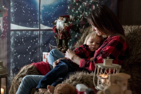 Beautiful young mother, hugging her toddler boy, sitting in cozy chair on Christmas, decoration around herの写真素材