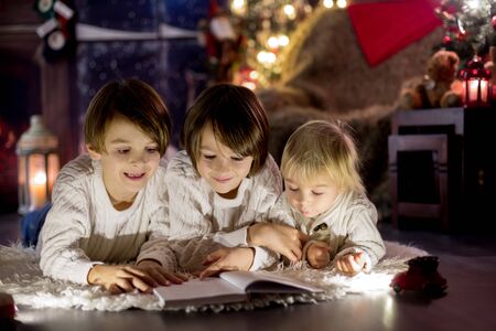 Three children, boy brothers, reading book at home at night on Christmas evening, lying on the floor