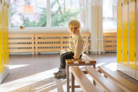 Sweet blonde toddler boy standing in front of a lockers in kindergarden or school hallway, wintertimeの写真素材