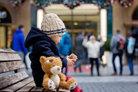 Fashion toddler boy with teddy bear toy, eating traditional Czech sweet pastry, called trdlo in the city center, sitting on bench,Christmas timeの写真素材