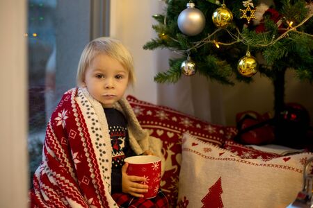 Sweet toddler boy in christmas pajama, sitting on window close to christmas tree, drinking milk and...