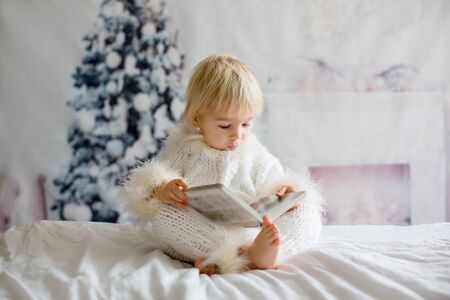 Blond toddler child, boy, reading a book at home, sitting in bed, dressed in fury overallの写真素材