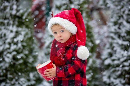 Sweet beautiful toddler boy, holding cup with hot milk, drinking outdoor in the snow, enjoying winter and Christmas holidaysの写真素材