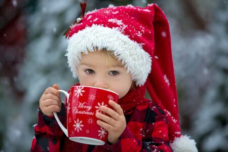 Sweet beautiful toddler boy, holding cup with hot milk, drinking outdoor in the snow, enjoying winter and Christmas holidaysの写真素材