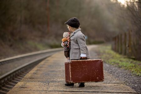 Adorable boy on a railway station, waiting for the train with suitcase and beautiful vintage porcelain dollの写真素材