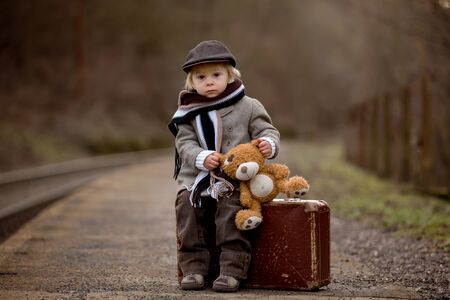 Adorable boy on a railway station, waiting for the train with suitcase and sweet teddy bear toyの写真素材