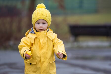 Beautiful funny blonde toddler boy with rubber ducks and colorful umbrella, jumping in puddles and playing in the rain, wintertimeの写真素材