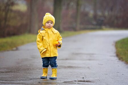 Beautiful funny blonde toddler boy with rubber ducks and colorful umbrella, jumping in puddles and...