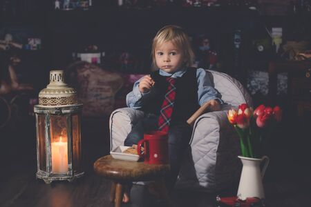 Sweet toddler boy, sitting in the dark, reading a book and drinking tea with cookies, sittng in a...