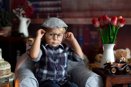 Sweet toddler boy, wearing glasses, reading a book and drinking tea with cookies, sittng in a cozy chairの写真素材