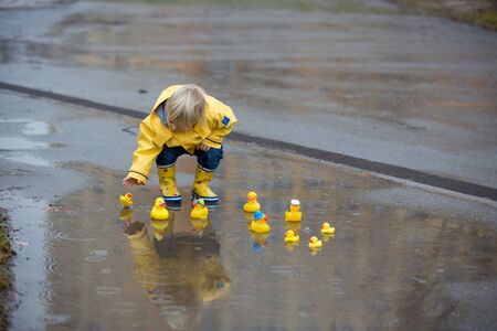 Beautiful funny blonde toddler boy with rubber ducks and colorful umbrella, jumping in puddles and...