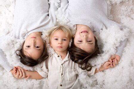 Beautiful portrait of three sibling boys, lying on the floor in a pile of cotton, holding hands and looking at cameraの写真素材