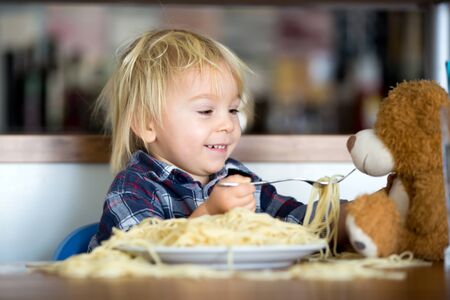 Little baby boy, toddler child, eating spaghetti for lunch and making feeding teddy bear friend, pot with spaghetti and tomatoes on the tableの写真素材