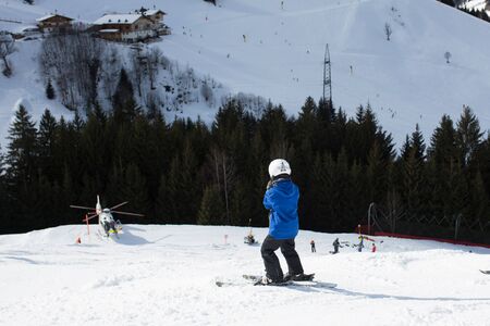 Child, watching rescue action on ski slope, helicopter picking up injured person from ski slope in...