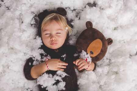 Cute blond toddler boy in knitted bear overall, holding teddy bear toy, sleeping in the snowの写真素材