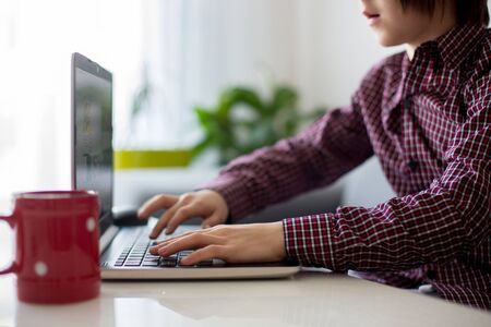 School child, sitting at the table with laptop, writing school tasks while homeschooling, while...