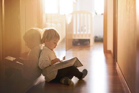 Beautiful toddler blond boy, lying on the floor at home in the hall, reading book, beautiful back lightの写真素材