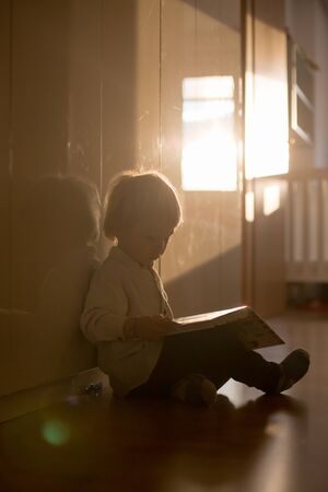 Beautiful toddler blond boy, lying on the floor at home in the hall, reading book, beautiful back lightの写真素材
