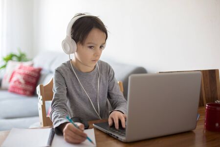 School child, sitting at the table with laptop, writing school tasks while homeschooling, while...