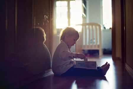 Beautiful toddler blond boy, lying on the floor at home in the hall, reading book, beautiful back lightの写真素材