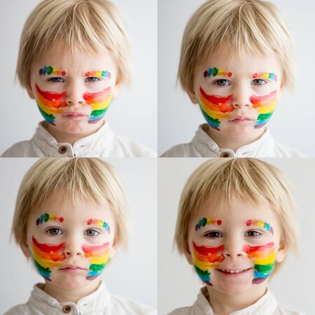 Beautiful blond toddler boy with rainbow painted on his face and messy hands, smiling happily