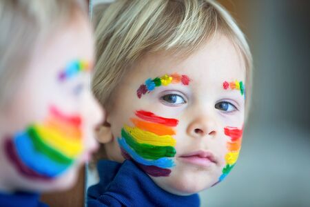 Beautiful blond toddler boy with rainbow painted on his face and messy hands, smiling happily