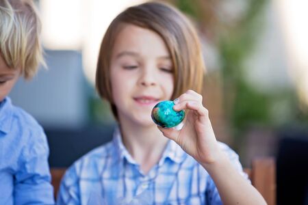Sweet children, brothers, coloring and paiting eggs for Easter in garden, outdoors at home in backyardの写真素材