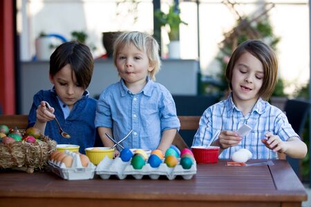 Sweet children, brothers, coloring and paiting eggs for Easter in garden, outdoors at home in...