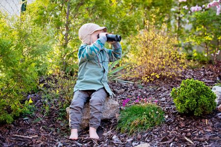 Blonde little toddler child with binoculars, sitting in garden on sunset, playing