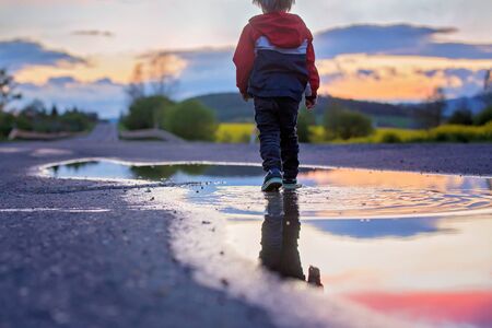 Sweet toddler child, cute boy with red raincoat, playing in puddle on sunsetの写真素材