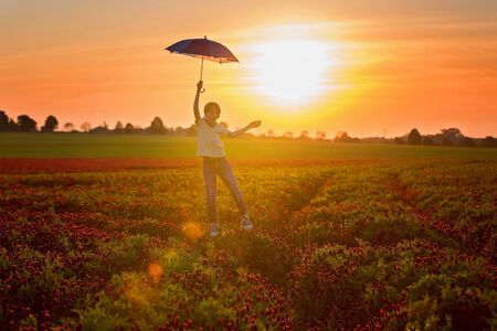 Beautiful child, flying with umbrella over gorgeous crimson clover field on sunset, springtimeの写真素材