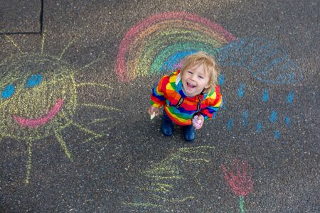 Cute toddler. playing in the rain with chalks, drawing on the asphalt and having funの写真素材