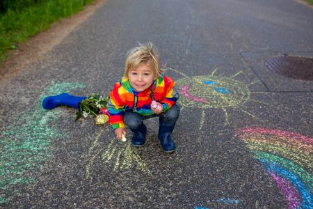 Cute toddler. playing in the rain with chalks, drawing on the asphalt and having funの写真素材