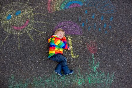 Cute toddler. playing in the rain with chalks, drawing on the asphalt and having funの写真素材