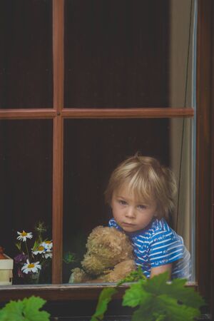 Sweet toddler boy, wearing medical mask, hugging teddy bear, also with mask, looking sadly out of the window, during coronavirus pandemic isolationの写真素材