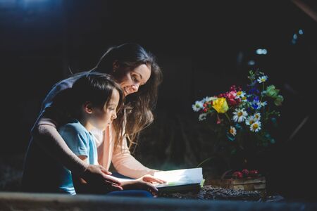 Beautiful mom and preschool boy, reading a book in the attic, nice atmosphere, flowers and strawberries next to himの写真素材