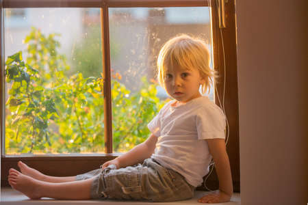 Abandoned little toddler boy, sitting sad on a window shield, looking outside at the sunsetの写真素材