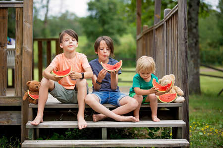 Three children, boy brothers, eating watermelon, sitting on stairs barefoot summertimeの写真素材
