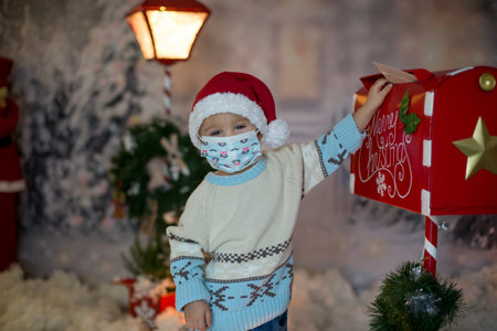 Little child, toddler boy, wearing medical mask, sending letter to santa in christmas mailbox, christmas decoration around him, outdoor snow shotの写真素材