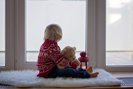 Sweet blonde child, boy, sitting on window shield with teddy bear friend toy, looking at the view outdoorsの写真素材