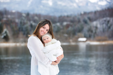 Beautiful mother in white dress and cute baby boy in knitted onesie, having taken their beautiful winter outdoor portrait on a sunny winter snowy dayの写真素材