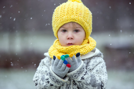 Beautiful blond toddler child, boy, with handmade knitted sweater playing in the park with first snow, enjoyingの写真素材