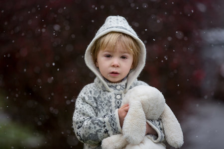 Beautiful blond toddler child, boy, with handmade knitted sweater playing in the park with first snow, enjoyingの写真素材