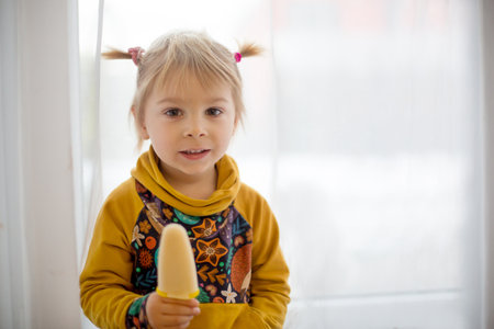Cute sweet child, toddler with little ponytails, goofying at home, eating ice cream, making funny faces and smilingの写真素材